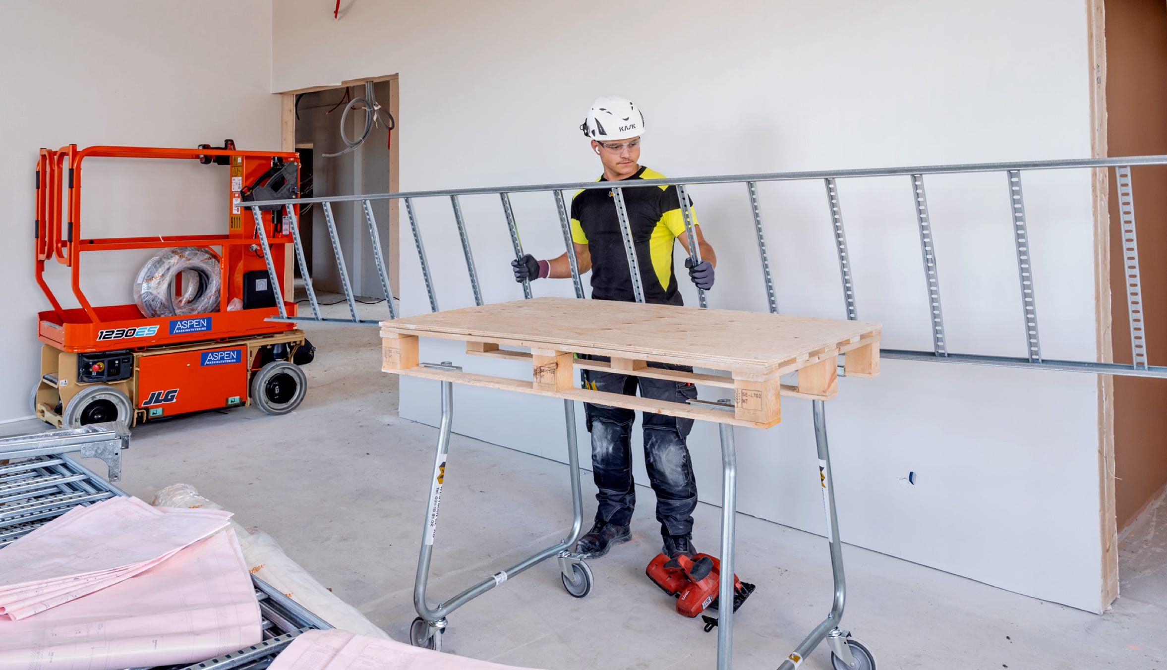 A person wearing a helmet and safety gear stands in a room with construction materials, including a large ladder and a wooden board on a cart. An orange lift is visible in the background.