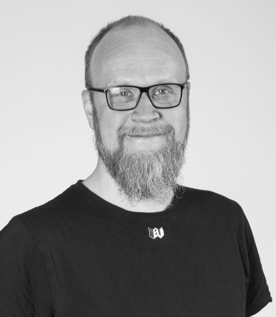 A black and white photo of a person with glasses, a beard, and a bald head, wearing a black t-shirt, smiling at the camera.