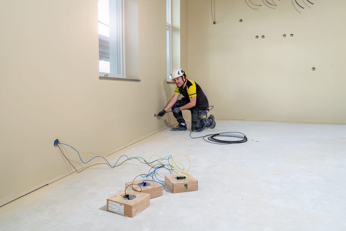 A person wearing a helmet and work clothes is kneeling on the floor of an empty room, working on electrical wiring near the wall. There are tools and cables on the floor.