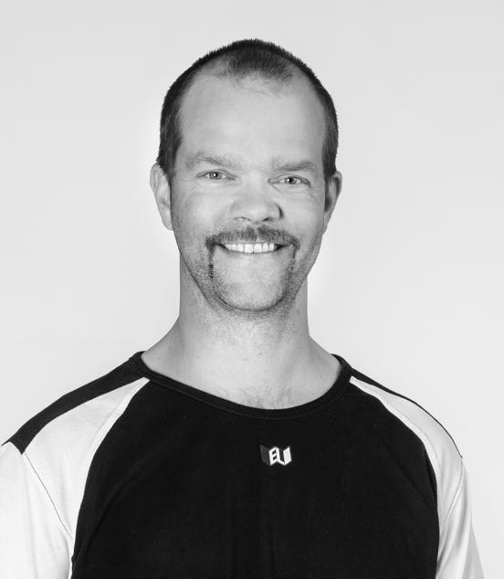 Man with a mustache smiling, wearing a black and white shirt, against a plain background.